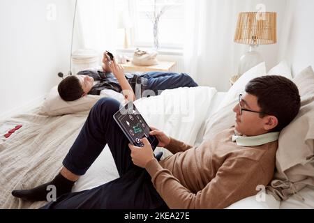 Brothers lying on bed and using phone and tablet Stock Photo