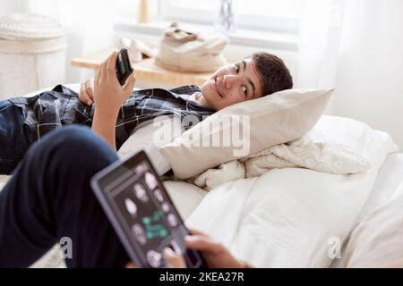 Brothers lying on bed and using phone and tablet Stock Photo