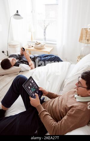 Brothers lying on bed and using phone and tablet Stock Photo