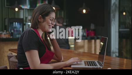 Female hispanic barista employee working in front of laptop computer taking online delivery. Young woman using technology speaking with client and mul Stock Photo