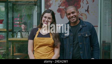 Portrait of two diverse young employees standing in front of coffee shop smiling. A female and male staff of small business. A young hispanic woman em Stock Photo