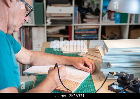 Senior man working with the pyrographer burning the wood making artwork craft in his art studio. Stock Photo