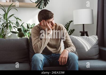 Unhappy male suffering from negative thoughts, having depressive mood or problems in relations. Regretting mistakes concept. Bored young man sitting o Stock Photo