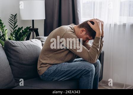 Upset young caucasian man sit on couch in living room at home and thinking, having heavy thoughts, financial difficulties concept. Stressed sad guy lo Stock Photo
