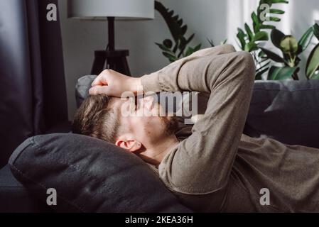Depression, apathy concept. Tired frustrated caucasian man 30s lying on sofa at home, thinking over problems, feeling stress, fatigue, touching head, Stock Photo