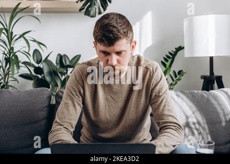 Close up of focused young man sitting on couch with laptop, working remotely online from home. Concentrated millennial male studying distantly alone i Stock Photo