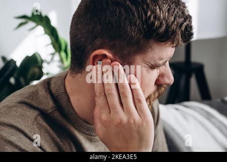 Close-up of unhealthy young caucasian man 30s touching ear, suffering from sudden throbbing ear ache, looking aside. Upset bearded male feeling unwell Stock Photo