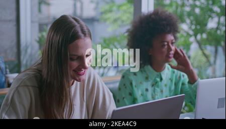 Two diverse young women at coffee shop in front of laptop computers. Female work colleagues working remotely Stock Photo