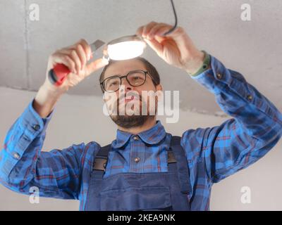 Electrician at work. Service for repair of electrical wiring and replacement of ceiling lamps. builder is installing small lamp on ceiling. Rent-a-gent helps with housework Stock Photo