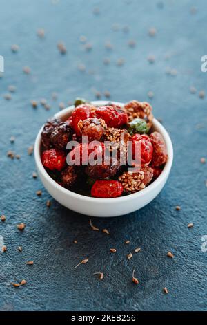 a white ceramic bowl with an assortment of candied peanuts, with different color sugar and seeds covers, placed on a dark gray stone surface Stock Photo