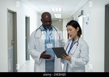 Two diverse female and female doctors looking at tablet and talking in hospital corridor Stock Photo