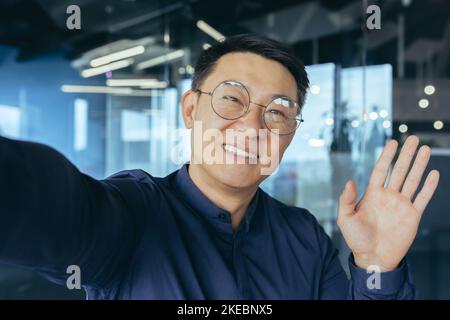 Smiling and cheerful asian businessman boss investor in glasses using smart phone for video call, looking into phone camera and waving hand gesture of greeting, talking with colleagues inside office. Stock Photo