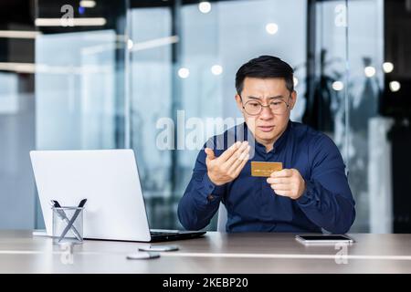 Upset and sad asian businessman inside office, man holding smartphone and bank credit card in hands, no funds in account received payment rejection, man working with laptop. Stock Photo