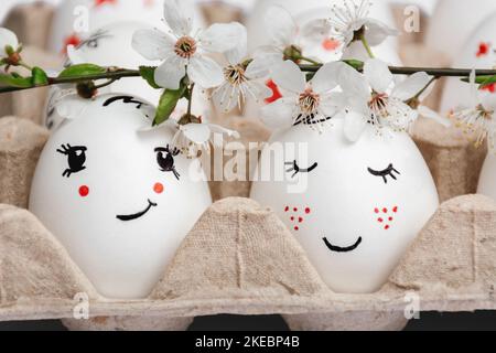 Close-up of delicately painted Easter eggs in paper eco-friendly tray. From above, sprig of spring white flowers from fruit trees. Pair of eggs looks like bride and groom. Spring style. Postcard Stock Photo