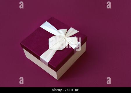 Soft selective focus on beautiful burgundy gift box with satin light ribbon. A festive gift for Christmas, New Year or romantic Valentine's Day. Copy space for text. Stock Photo