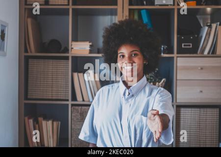 Portrait of attractive dark skinned young woman with curly Afro hairstyle extending hand for handshake. African american businesswoman smiling at office. Young positive lady reaching out to greet someone Stock Photo