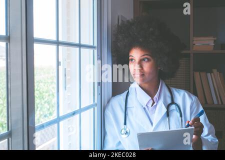 Thoughtful female african american doctor with stethoscope, documents and pen looking outside her clinic window. Serious contemplative frontline heathcare worker admiring view from window Stock Photo