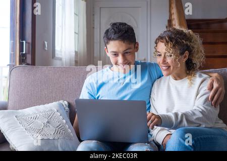 Adult son and middle-aged 40s mother sitting on couch in cozy living room holding on lap notebook laughing on funny comedy movie, enjoy on-line amusements websites, internet virtual fun online concept Stock Photo