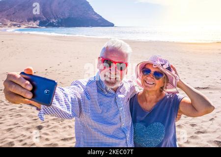 couple of mature people or pensioners enjoying their vacations and summer time together on the sand of the beach with the sea or ocean at the background - two retired senior taking a selfie looking at the phone Stock Photo