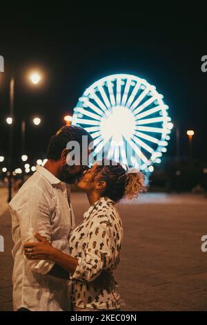 Loving couple embracing and kissing each other passionately on street against illuminated ferris wheel at night. Couple romancing and spending quality time outdoors during vacation at night Stock Photo
