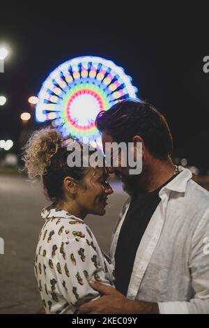 Loving couple embracing each other passionately on street against illuminated ferris wheel at night. Couple romancing and spending quality time outdoors during vacation at night Stock Photo