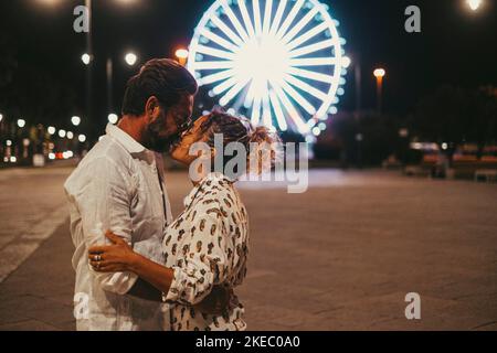 Loving couple embracing and kissing each other passionately on street against illuminated ferris wheel at night. Couple romancing and spending quality time outdoors during vacation at night Stock Photo
