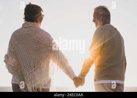 couple of two happy and active seniors having fun and enjoying together summer at the beach walking holding their hands with the sunset at the background Stock Photo