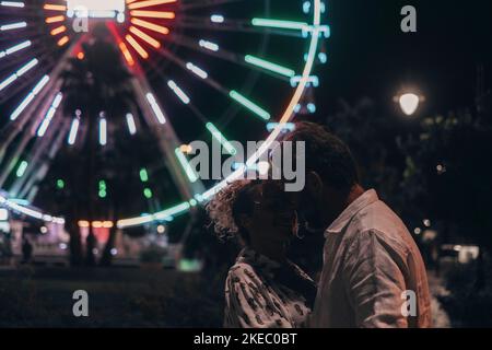 Loving couple embracing each other passionately on street against illuminated ferris wheel at night. Couple romancing and spending quality time outdoors during vacation at night Stock Photo