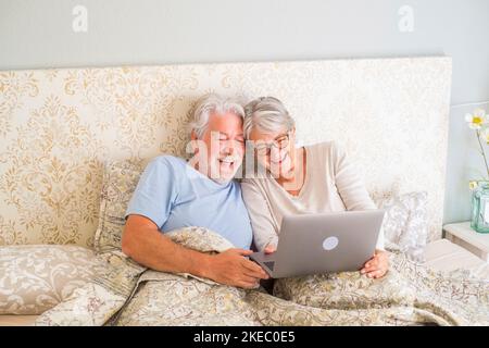 Old senior caucasian couple laughing and using laptop in the morning at bed in the bedroom at home. Elderly couple surfing and using social media on laptop at home. Stock Photo