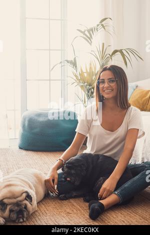 Portrait of beautiful woman in eyeglasses having fun with her pets pugs dogs sitting on floor in the living room of her house. Cheerful woman spending leisure time with her two cute pets dog at home Stock Photo