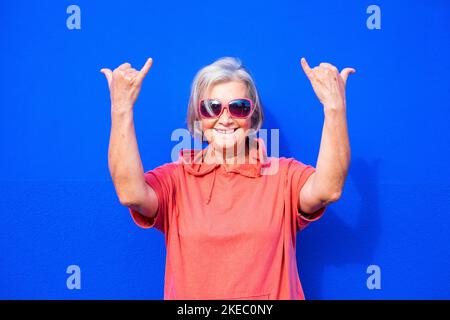 close up and portrait of funny and crazy senior or mature woman doing surfing sign and smililing looking at the camera with blue background - pensioner wearing red clothes and sunglasses happy people Stock Photo