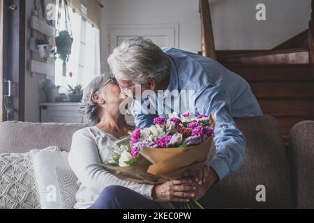 Old man giving flowers at his wife sitting on the sofa at home for the San Valentinesí day. Pensioners enjoying surprise kissing together. In love people having fun. Stock Photo