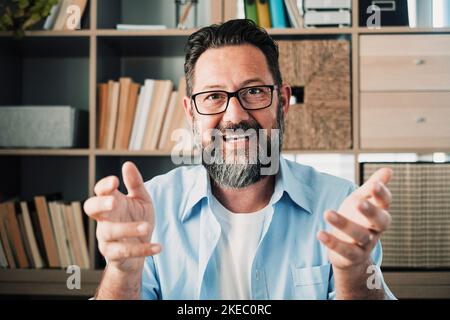 Head shot portrait confident businessman coach wearing glasses looking at camera and talking, mentor speaker holding online lesson, explaining, sitting at wooden work desk in modern cabinet Stock Photo