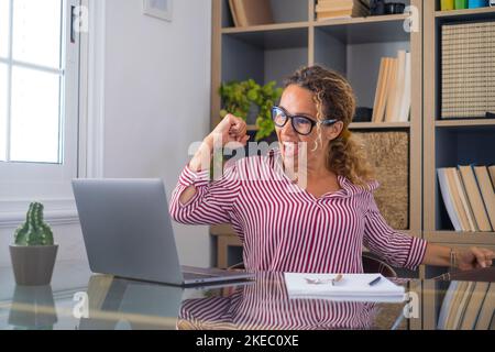 Caucasian woman office worker feeling excited raising fist celebrating career promotion or reward. Businesswoman delighted on receiving good news online using laptop at office workplace Stock Photo