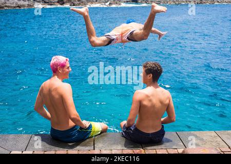 group of three teenager having fun and enjoying together summer and sea or ocea at the beach - two men boys sitting talking while one of them is jumping on their heads to the water - vacations lifestyle and concept Stock Photo