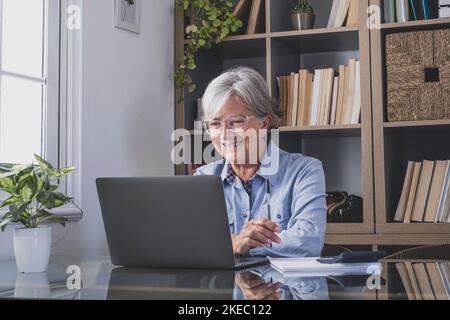 Happy old caucasian businesswoman smiling working online watching webinar podcast on laptop and learning education course conference calling make notes sit at work desk, elearning concept Stock Photo