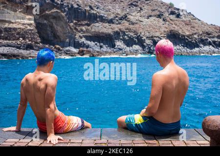two happy teenagers together at the beach having fun sitting on the floor - two people wiwht crazy and colored hair talking and enjoying summer at the beach with the sea Stock Photo