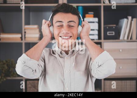 Tranquil carefree young teenager man at home, wearing modern wireless headphones, listening to favorite classic music online, feeling peaceful mindful alone in living room. Stock Photo