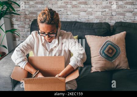 Woman unpacking delivdery box opening package at home. Happy young lady looking at carton box while sitting on sofa in living room at modern apartment. Caucasian female checking out delivered stuff. Stock Photo