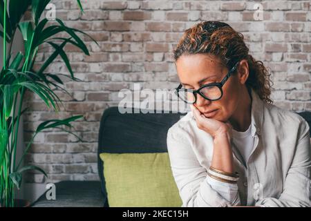 Thoughtful sad young woman in casual clothes sitting on couch in cozy modern living room looking down at home. Depressed caucasian female in deep thoughts feeling lonely at her house Stock Photo