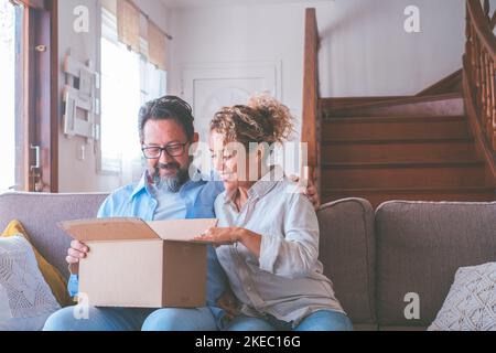 Happy caucasian couple looking inside cardboard box, satisfied with ordered item. Husband and wife sitting on sofa opening carton parcel in living room of house. Man and woman clients unbox package with internet order Stock Photo