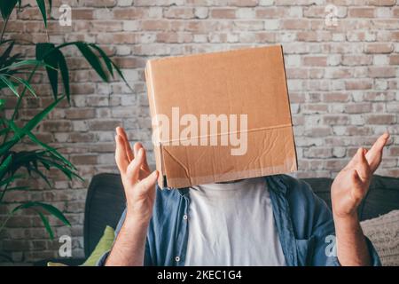 Mature man wearing empty carton box sitting on sofa and gesturing in the living room of house. Caucasian guy with face hidden or covered in cardboard delivery container while relaxing on couch at home Stock Photo