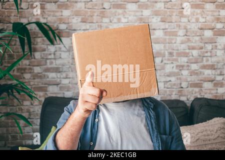 Mature man wearing empty carton box sitting on sofa and pointing finger in the living room of house. Caucasian guy with face hidden or covered in cardboard delivery container while relaxing on couch at home Stock Photo
