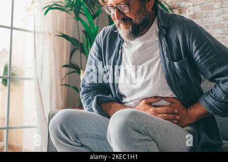 Man suffering with severe stomach pain sitting at home. Hand of mature guy holding abdomen suffering from ache, diarrhea or indigestive problem. Caucasian guy pressing on belly on painful sensation Stock Photo