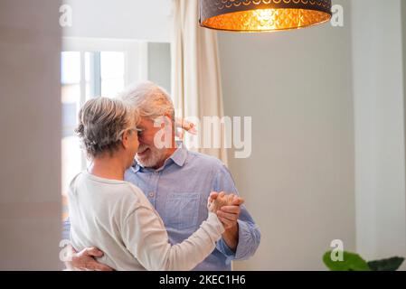 Cheerful senior couple dancing and smiling looking at each other. Elderly happy couple celebrating by dancing in living room. Romantic old aged couple holding hands and dancing together at home Stock Photo