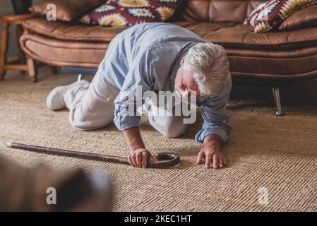 Elder senior man lying on floor after falling down with wooden walking stick beside couch on rug in living room at home. Old man suffering with pain and struggling to get up after falling down at house Stock Photo