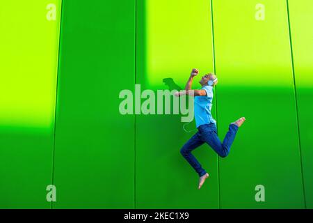 portrait and close up of teenager or millennial or young man jumping having fun and listening music with headphones - green background wall - blonde hair trendy concept - happy man enjoying lifestyle and concept Stock Photo