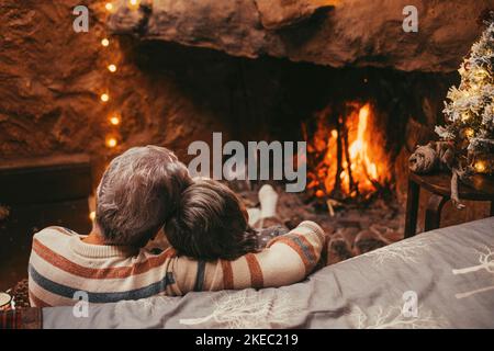 Old caucasian couple spending leisure time together at home. Loving romantic husband and wife relaxing while looking at burning fireplace during winter christmas holiday Stock Photo