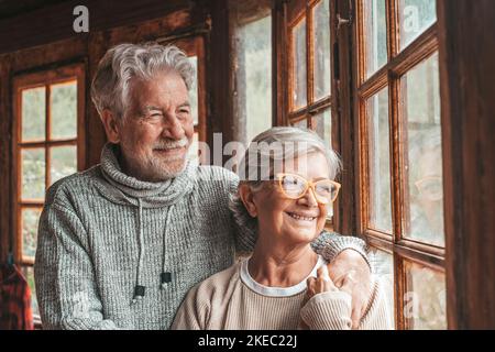 Old caucasian couple spending leisure time looking out through window at home. Loving husband embracing wife from behind and admiring something interesting from transparent door glass. Stock Photo