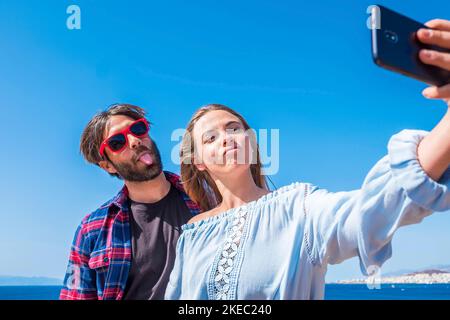 two happy and crazy people taking a selfie on the beach together doing crazy facial expressions and faces - couple of adults enjoying their trip outdoor - technology and social media lifestyle and concept Stock Photo